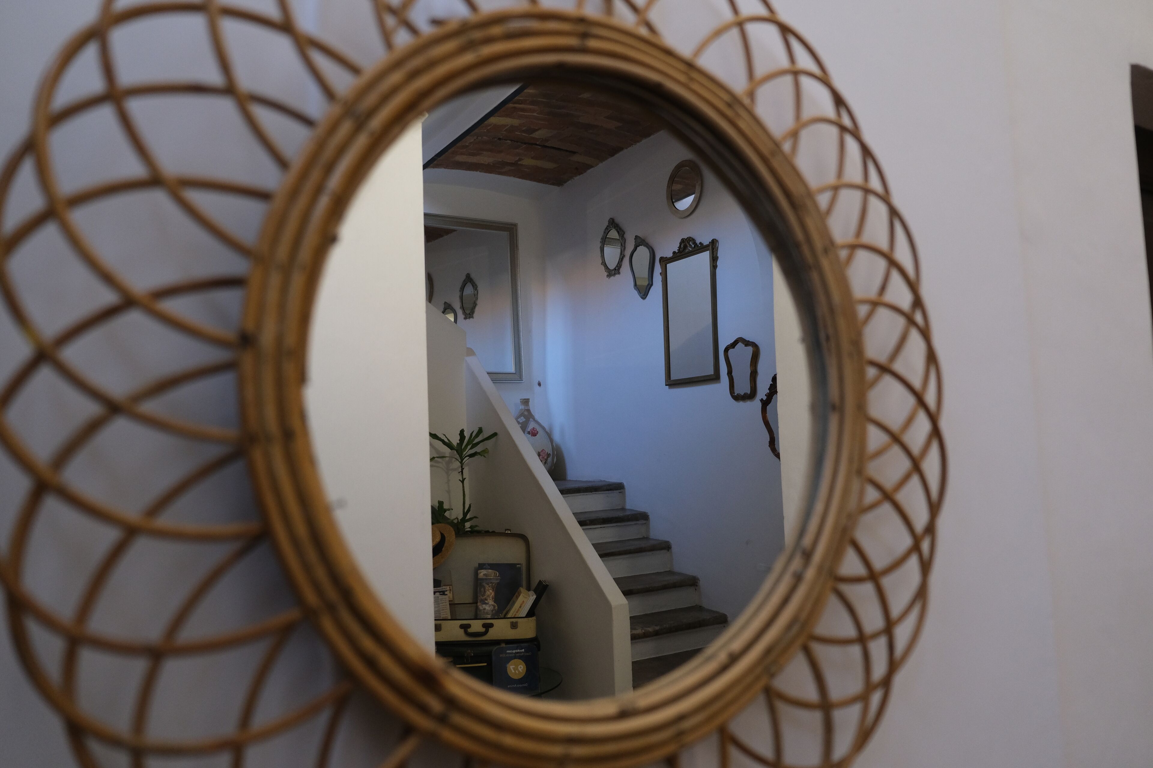 A round mirror with a geometric frame, reflecting a staircase and a small table with a plant and a suitcase.