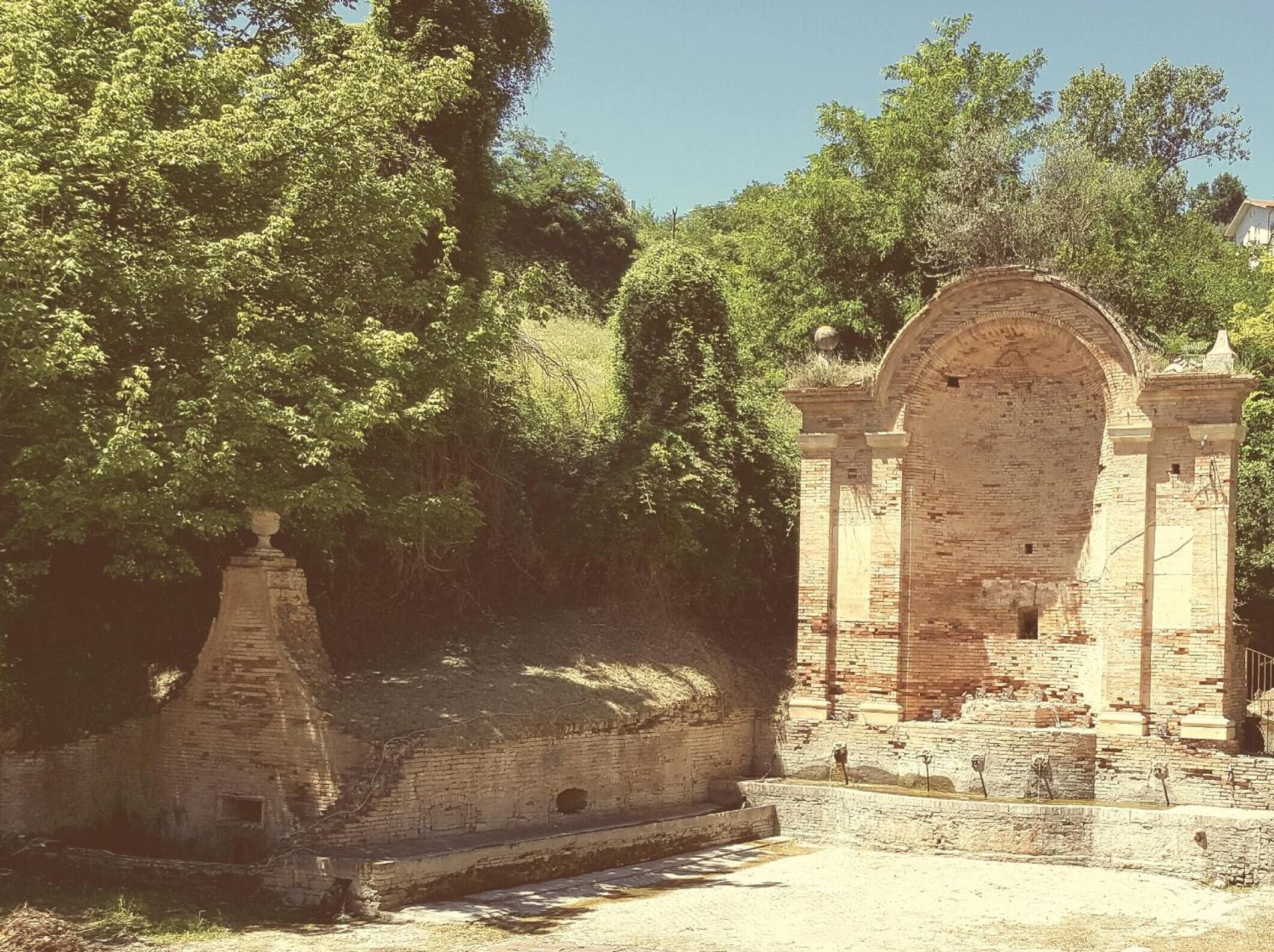 An ancient stone structure with a large arched gateway, surrounded by trees and a small pathway.