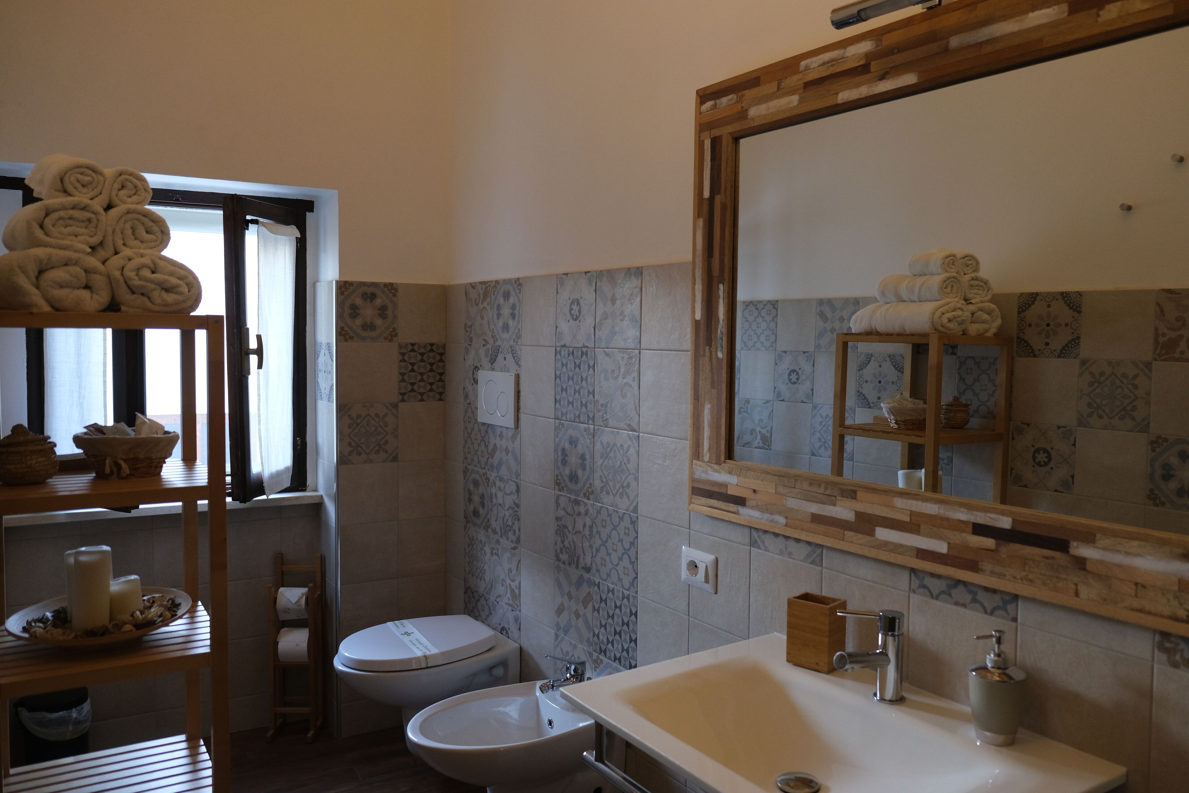 A bathroom with a sink, toilet, and a mirror with a wooden frame. There are towels stacked on a shelf and a window with curtains.
