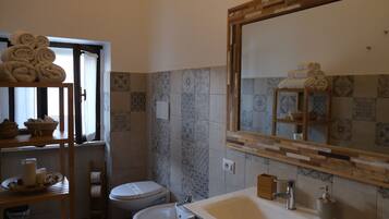 A bathroom with a sink, toilet, and a mirror with a wooden frame. There are towels stacked on a shelf and a window with curtains.