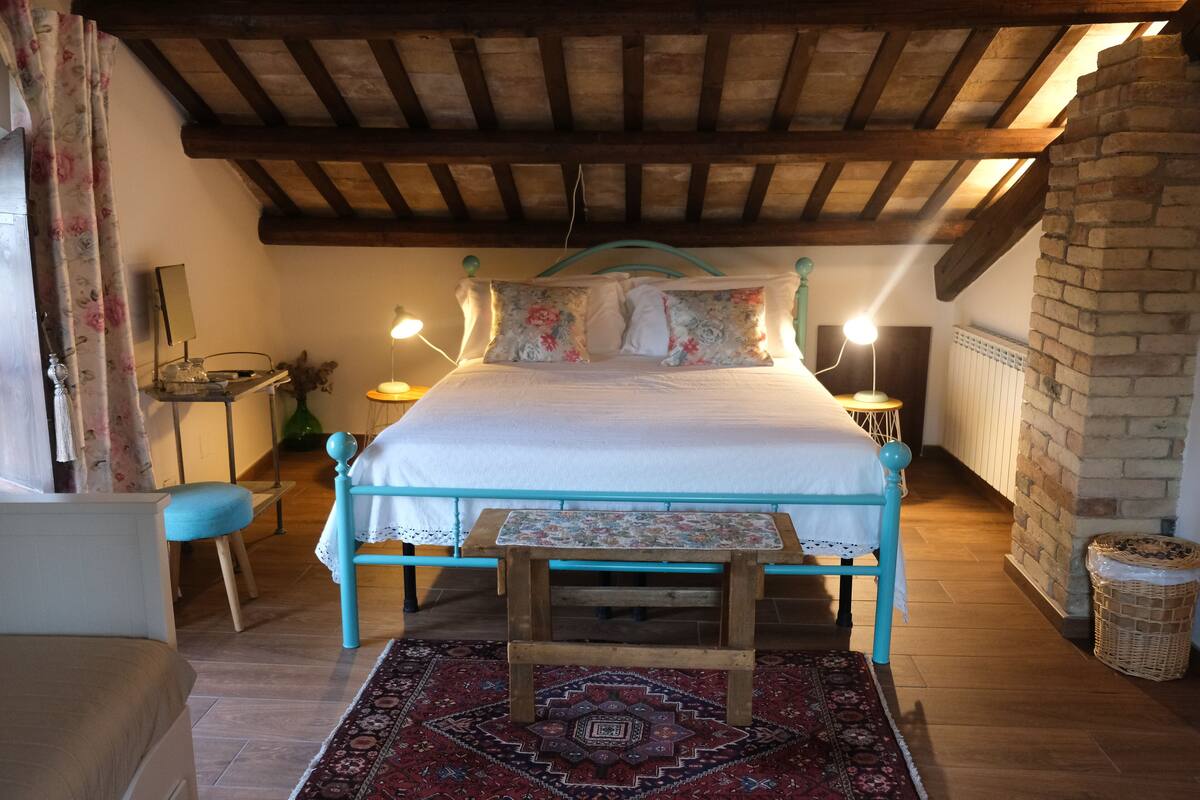 A bedroom with a wooden bed frame, a matching bench, a patterned rug, and a brick wall.