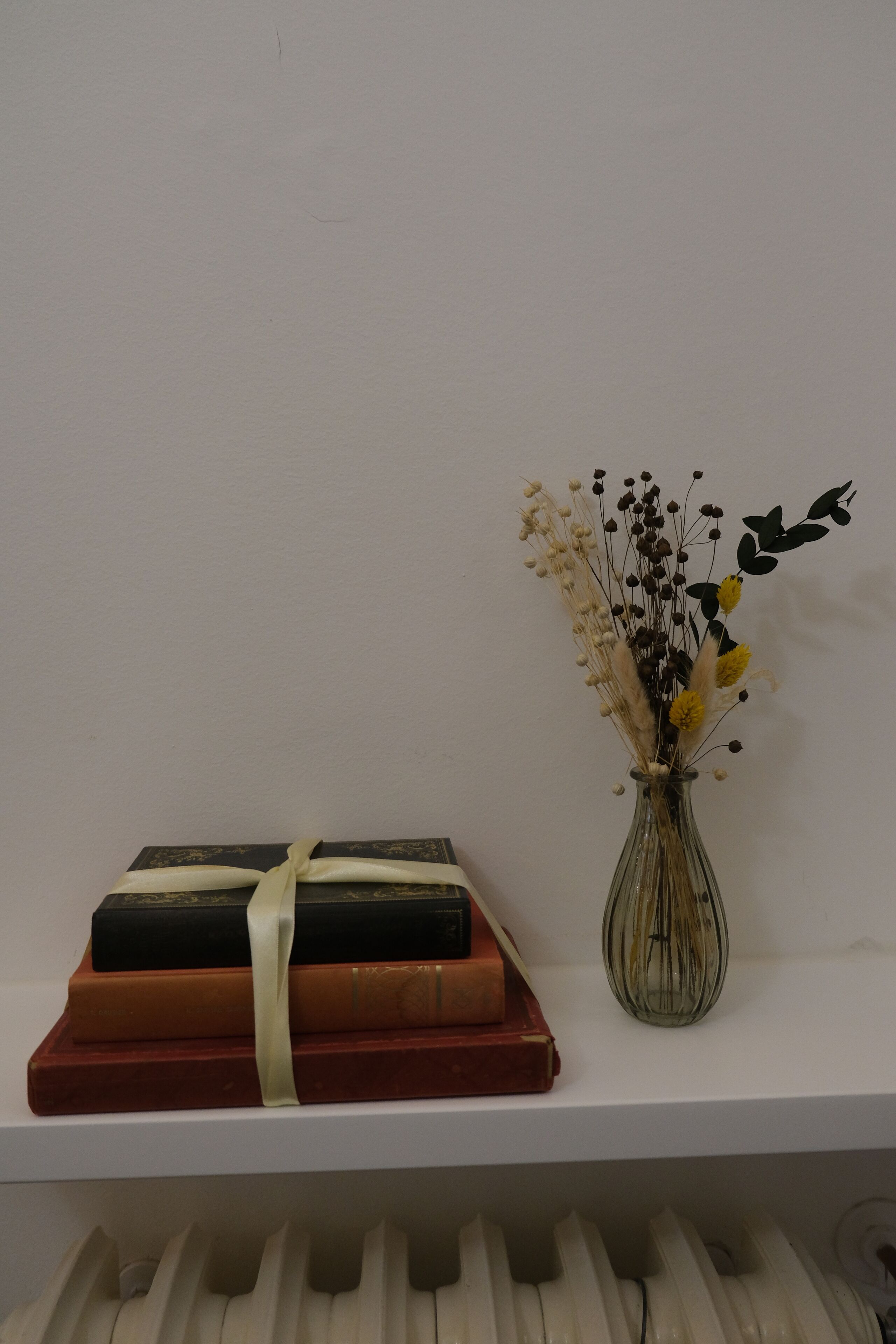 Una estantería blanca con una pila de libros y un jarrón de flores secas.