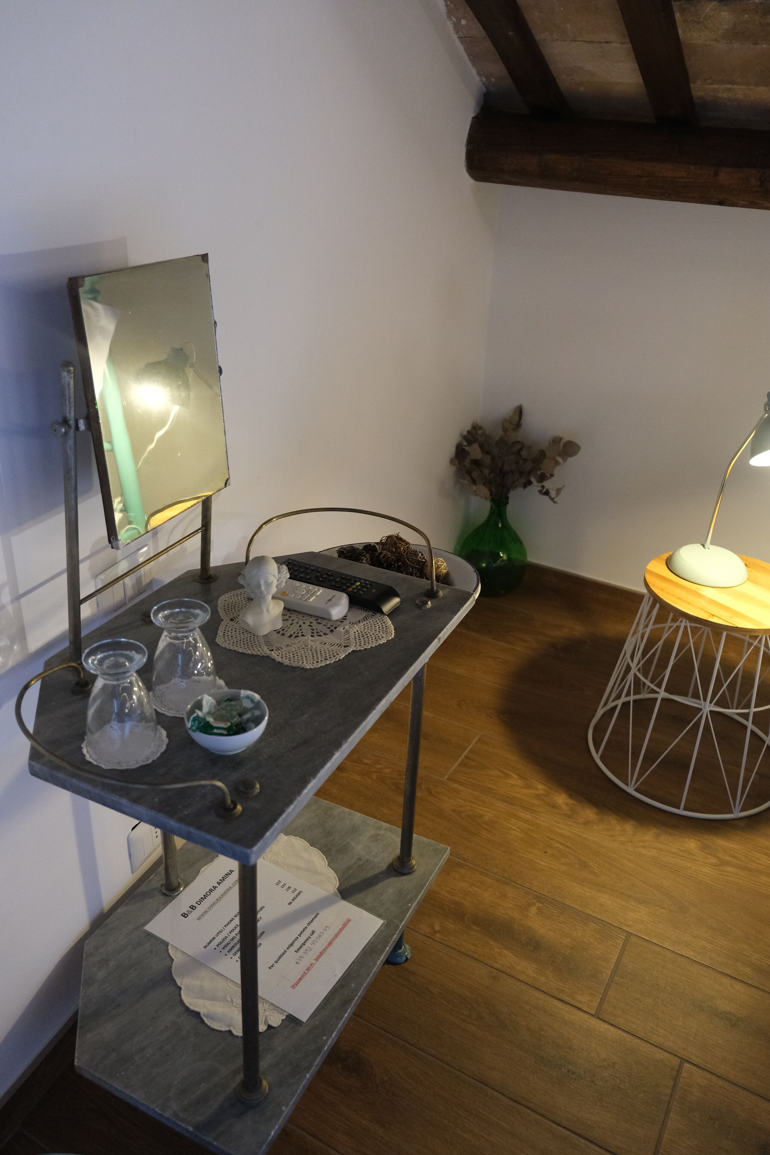 A room with a wooden floor, a metal table with a vase and a remote control, a small white table with papers, and a green vase with dried flowers.