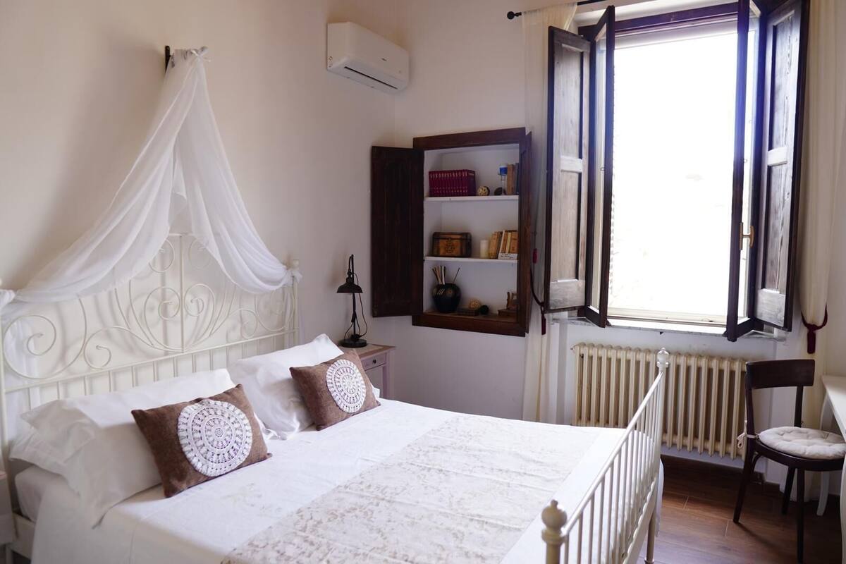 A bedroom with a four-poster bed, a bedside table with a lamp, a shelf with books and decorative items, and a window with curtains.