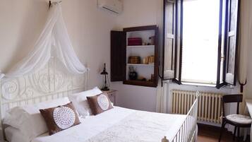 A bedroom with a four-poster bed, a bedside table with a lamp, a shelf with books and decorative items, and a window with curtains.