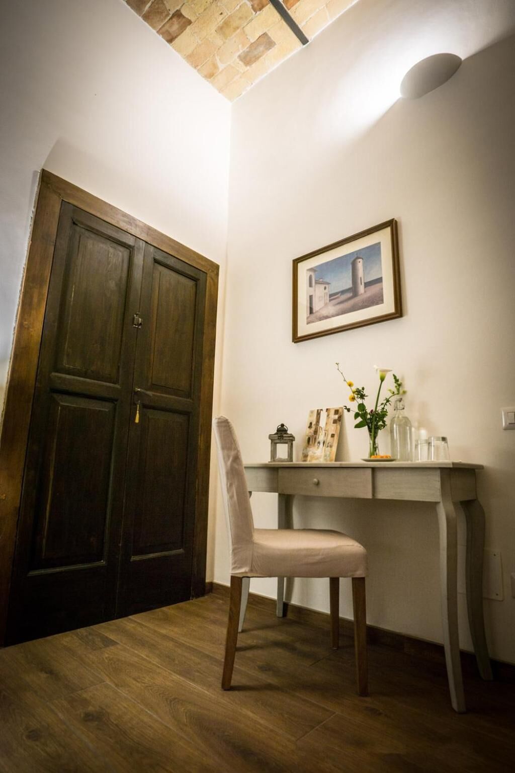 Ein Zimmer mit Holztür, Schreibtisch mit Stuhl, einem gerahmten Bild an der Wand und einer Vase mit Blumen.