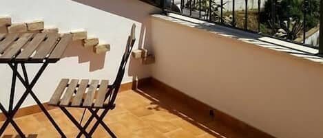 Un balcón con mesa y sillas de madera, un toldo blanco y vistas a un jardín y un edificio al fondo.