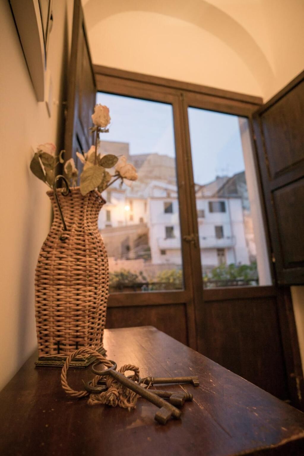 En fletkurv med tørrede blomster på et træbord, et vindue med udsigt til bygninger, og en dør i baggrunden.