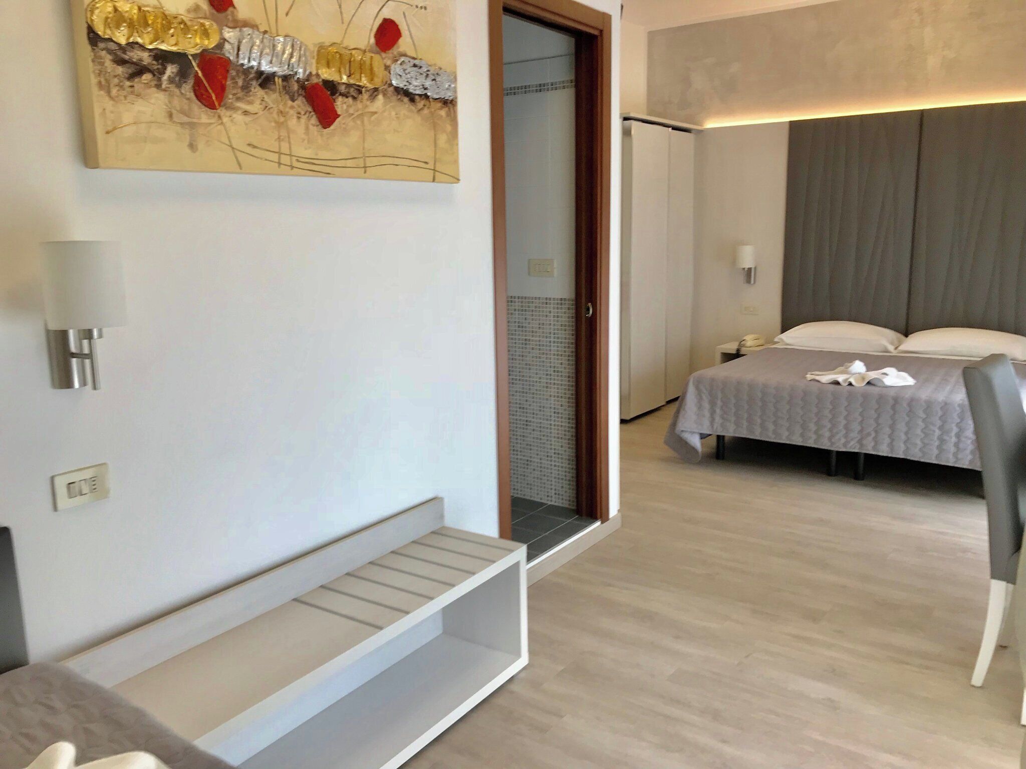 A hotel room with a bed, a chair, a small table, a wall-mounted light, and a door leading to a bathroom.