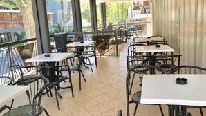 An indoor dining area with tables and chairs, large windows, and a view of greenery outside. - Hotel Bremen  (Rimini)