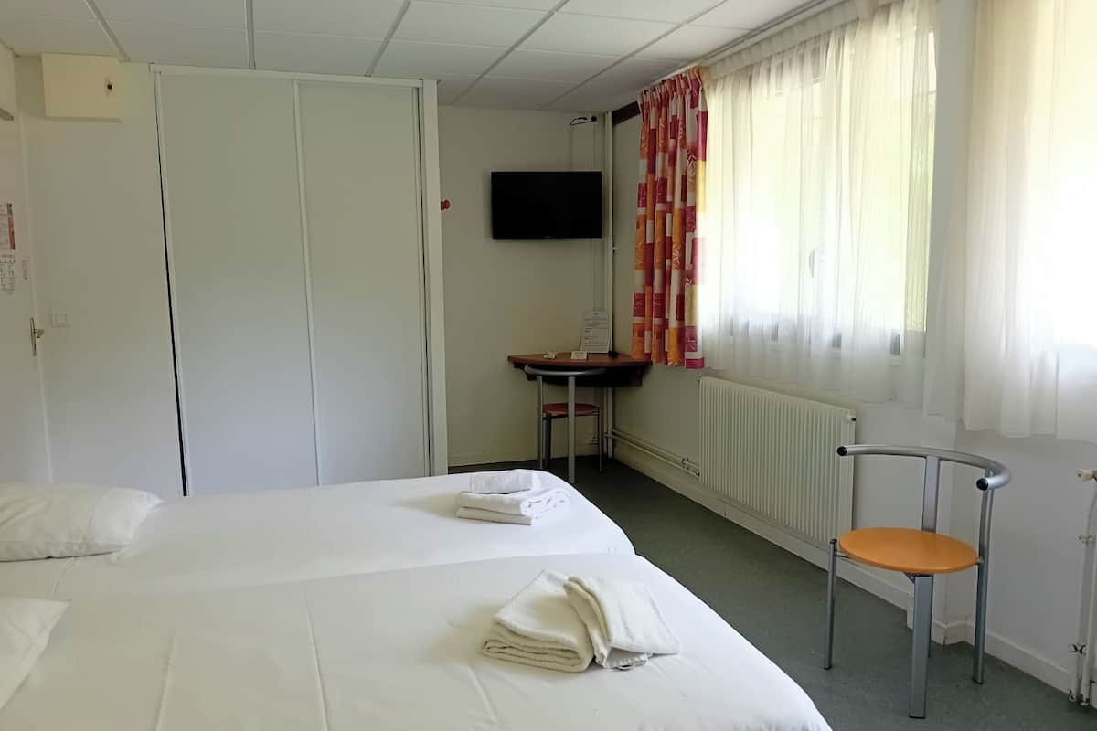 Ein Hotelzimmer mit zwei Betten, einem Schreibtisch, einem Stuhl, einem Fernseher und einem Fenster mit Vorhängen.