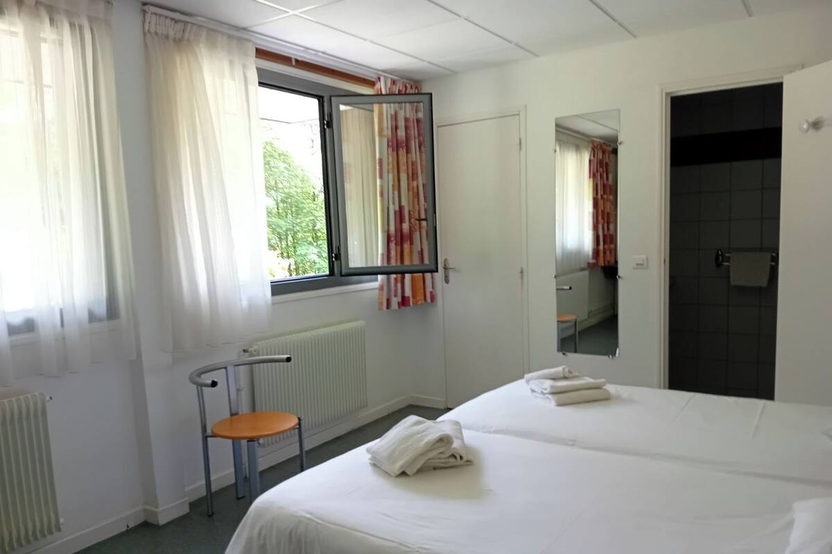 Ein Hotelzimmer mit zwei Betten, einem Fenster mit Vorhängen, einem Heizkörper, einem kleinen Tisch und einem Spiegel an der Wand.