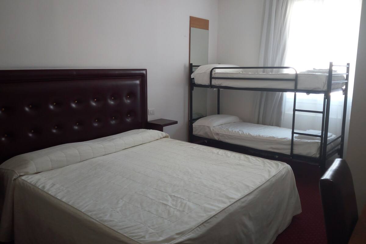 Ein Hotelzimmer mit einem Einzelbett, einem Etagenbett, einem hölzernen Kopfteil, einem Nachttisch, einem Spiegel und einem Fenster mit Vorhängen.