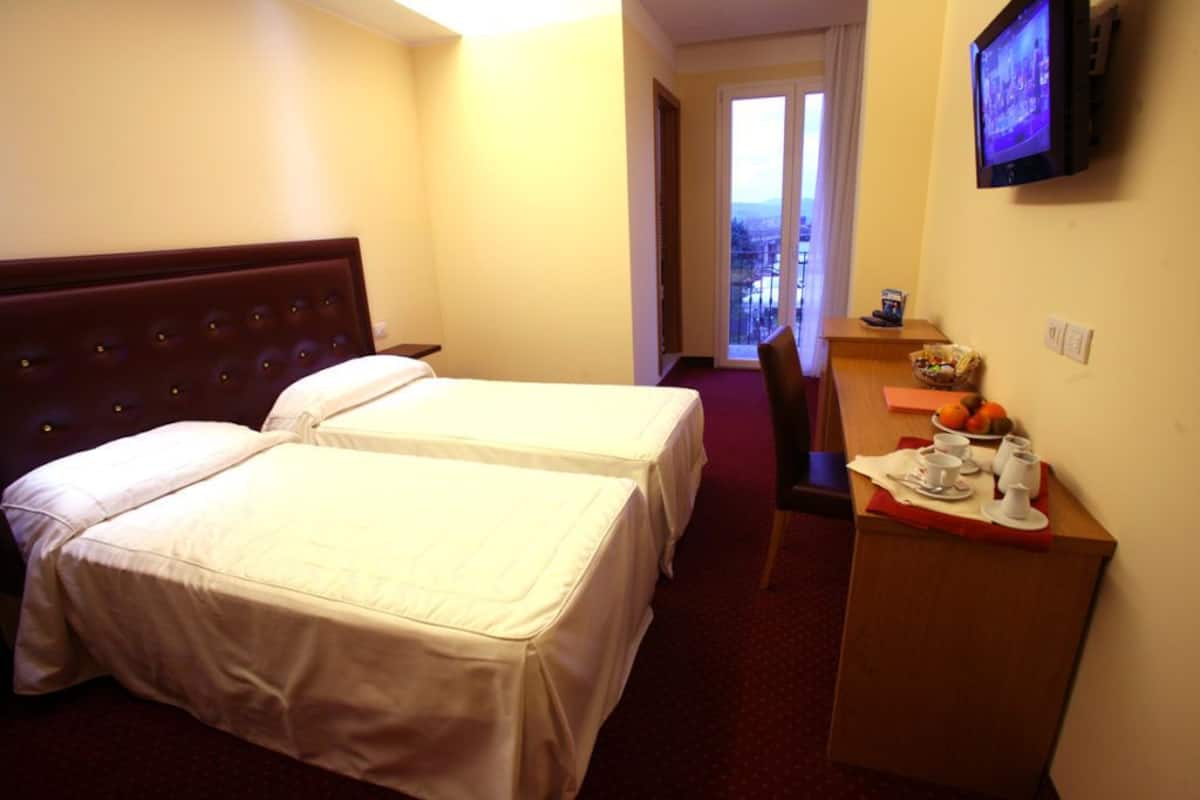 Ein Hotelzimmer mit zwei Betten, einem Schreibtisch mit Frühstücksangeboten, einem Fernseher und Blick auf die Stadt durch das Fenster.