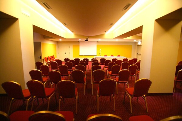 A conference room with rows of red chairs, a white projection screen, and a yellow accent wall. - Hotel Il Perugino (Corciano)