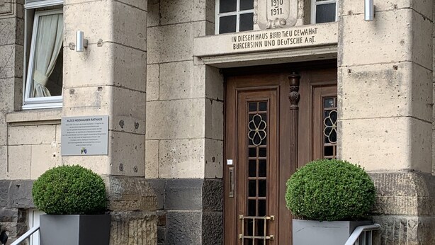 A stone building with a wooden door, two potted plants, and a sign indicating the building's historical significance. - Hotel Rathaus 1910 (Essen)