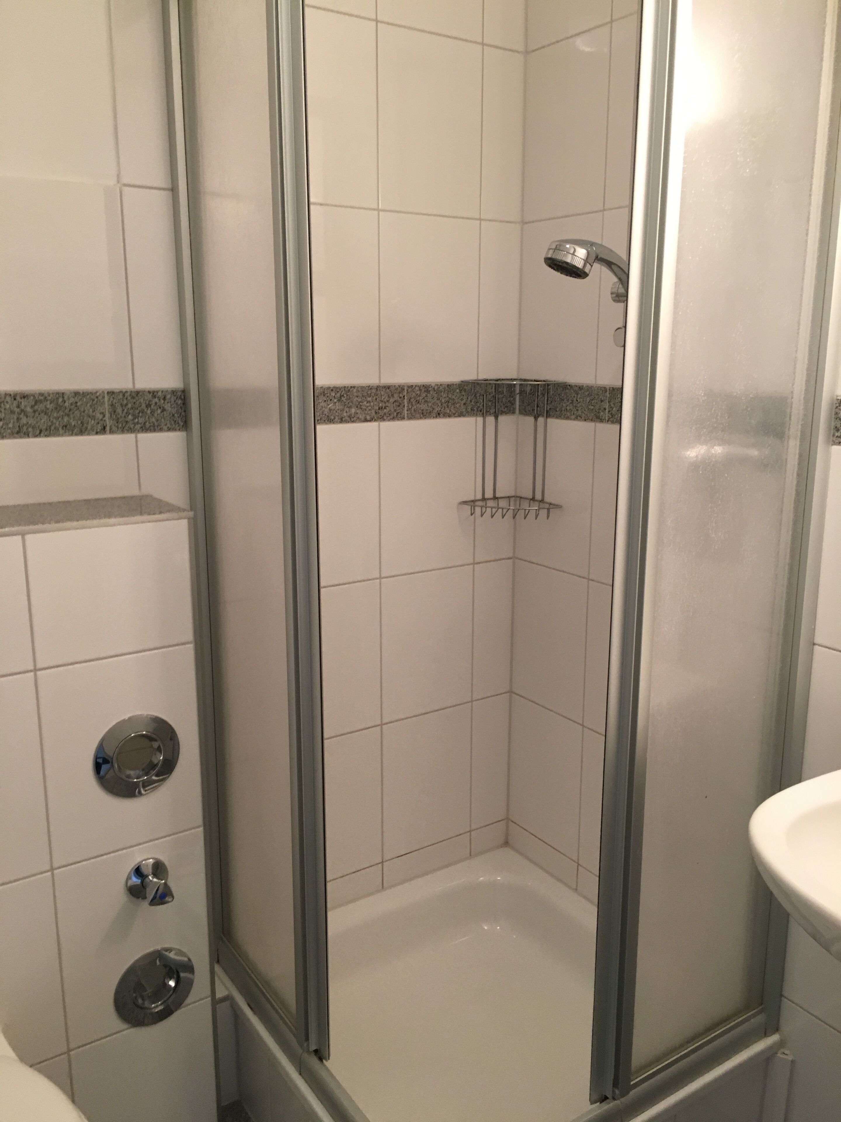 Eine Dusche mit Glaswand, weißen Fliesen und einer Metallbrause.
