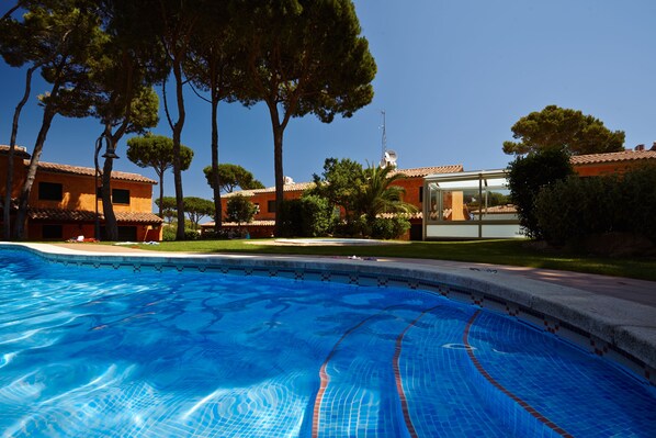 A swimming pool with clear blue water, surrounded by trees and buildings with orange walls and tiled roofs.
