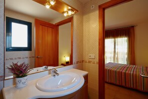 A bathroom with a white sink, a mirror, a wooden door, and a small bedside table with a lamp.