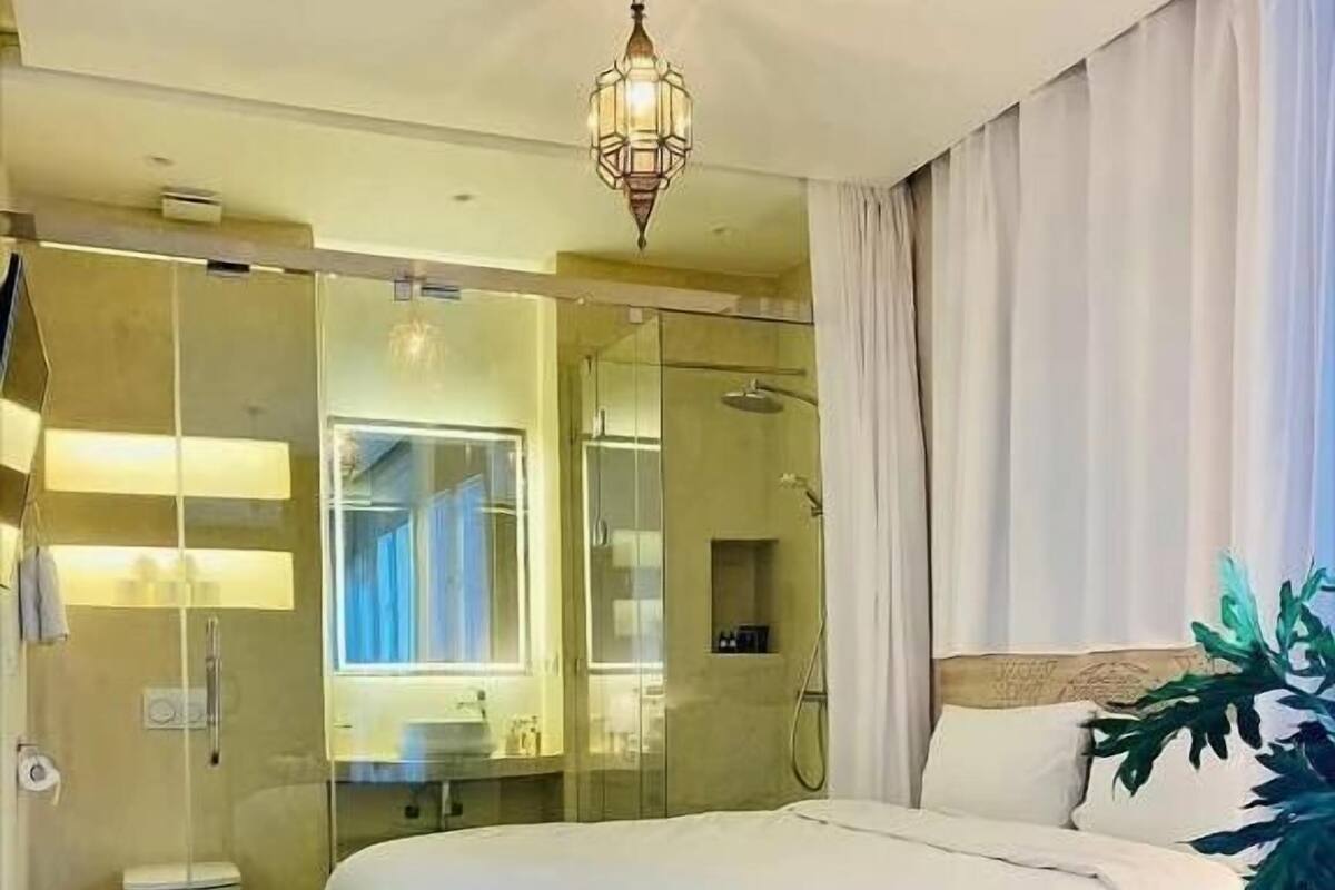 Hotellihuone, jossa on suuri sänky, lasiseinällä varustettu suihku, yöpöytä ja riippuvalaisin.