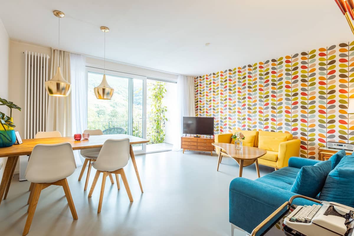 Un salon moderne comprenant un coin repas, un téléviseur et un mur décoré d’un motif coloré représentant des feuilles.