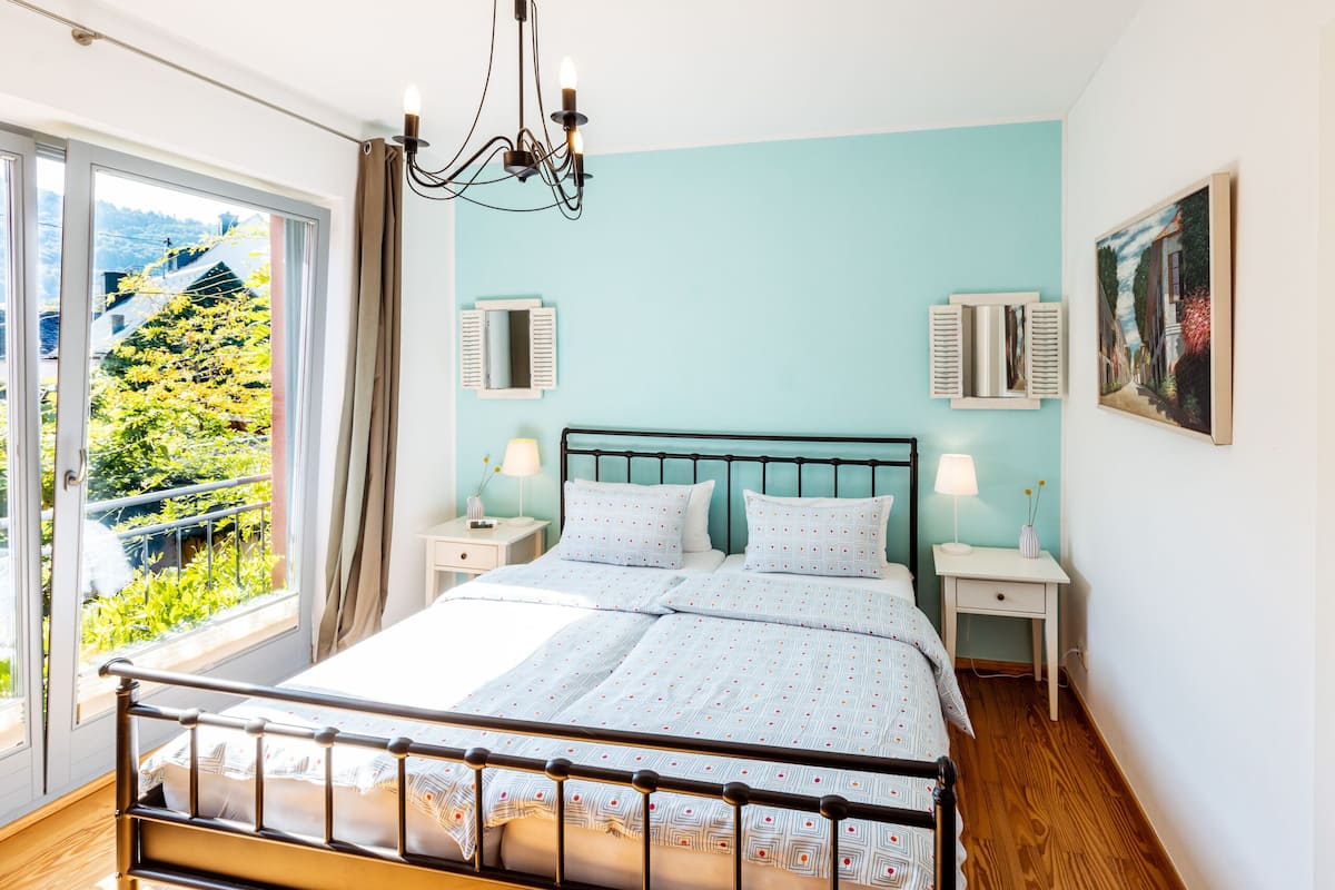 Une chambre avec un grand lit, deux tables de chevet, un lustre et une vue sur la verdure.