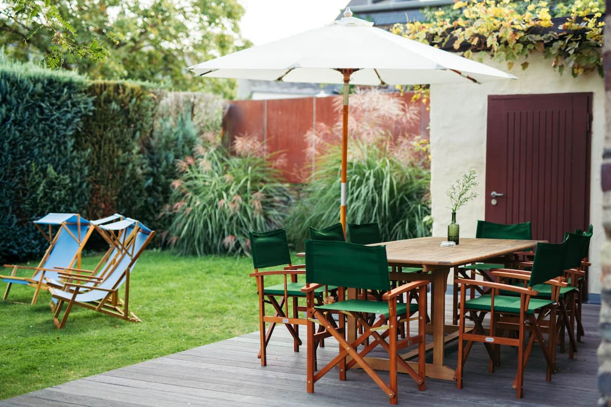 Une terrasse extérieure avec une table et des chaises en bois, entourée d’un jardin et d’une porte rouge.