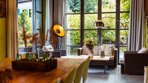 Un comedor acogedor con una mesa de madera, sillas de mimbre y un sofá con cojines. Hay un ventanal con vistas al follaje exterior.