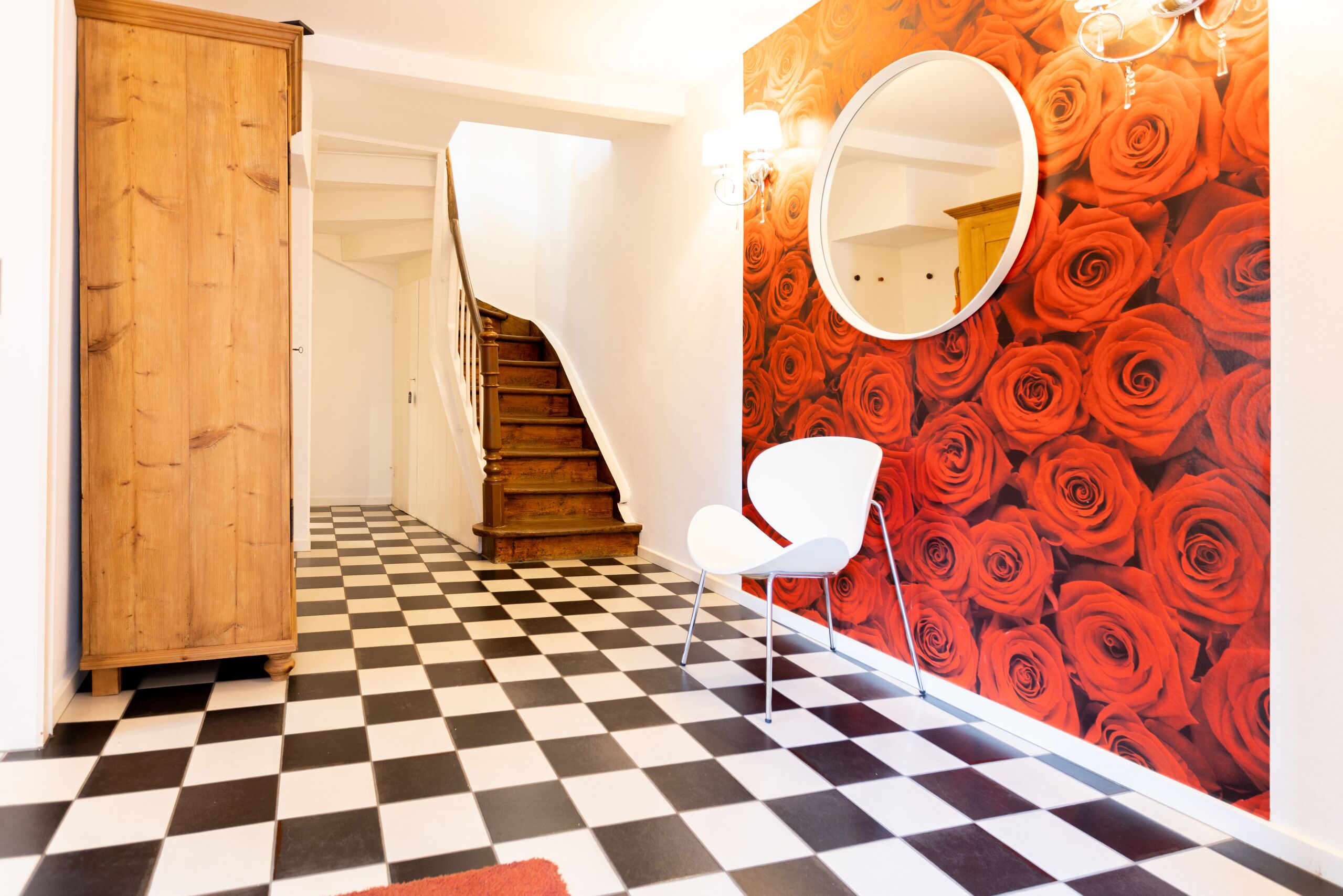 Un pasillo con piso a cuadros blanco y negro, un armario de madera, una silla blanca, un espejo redondo y una pared con estampado de rosas rojas.