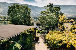 Um jardim com um caminho pavimentado, um edifício com telhado inclinado e vista para um lago e colinas ao fundo.