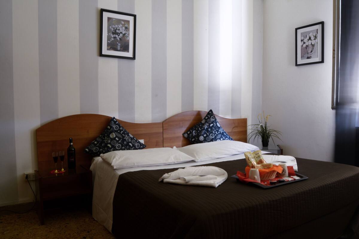 A hotel room with a bed, pillows, a tray with snacks, a bottle, and glasses.