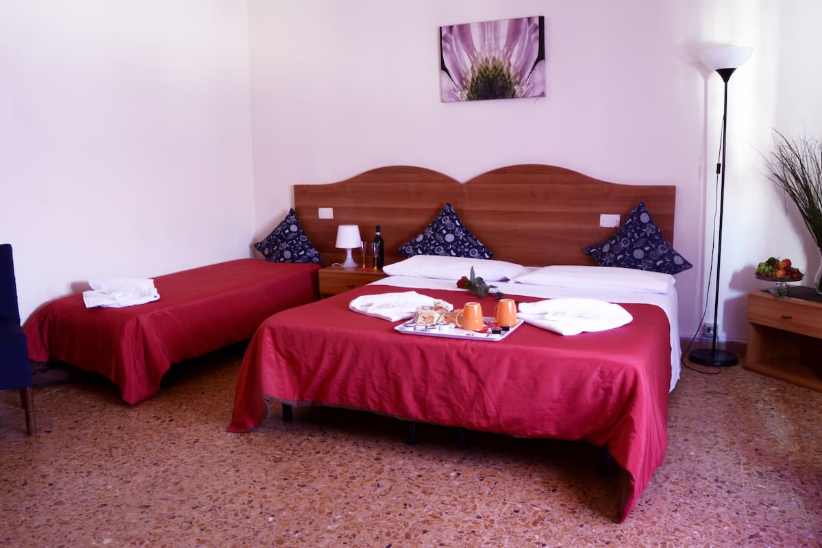 A hotel room with two single beds, each with a red bedspread and matching pillows. There is a bedside table with a lamp, a bottle, and a glass. The room has a tiled floor and a painting on the wall.