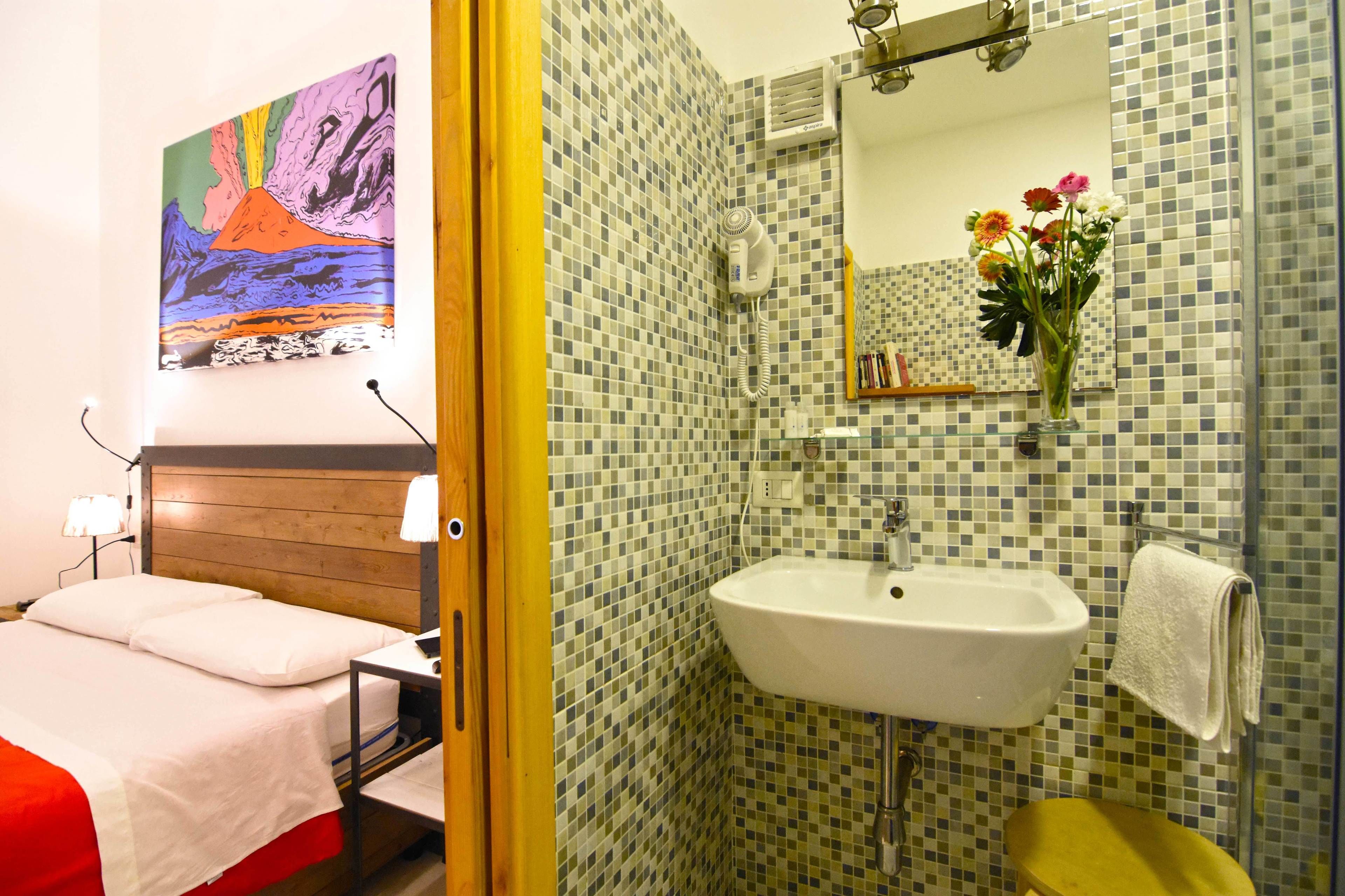 A hotel room with a bed, a bedside table, a nightstand, a wall-mounted painting, a bathroom with a sink, a mirror, a towel rack, and a shower area.