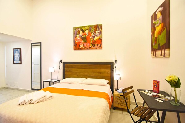 Une chambre d’hôtel avec un lit, une table de chevet, une table ornée d’un vase de fleurs et deux tableaux accrochés au mur.