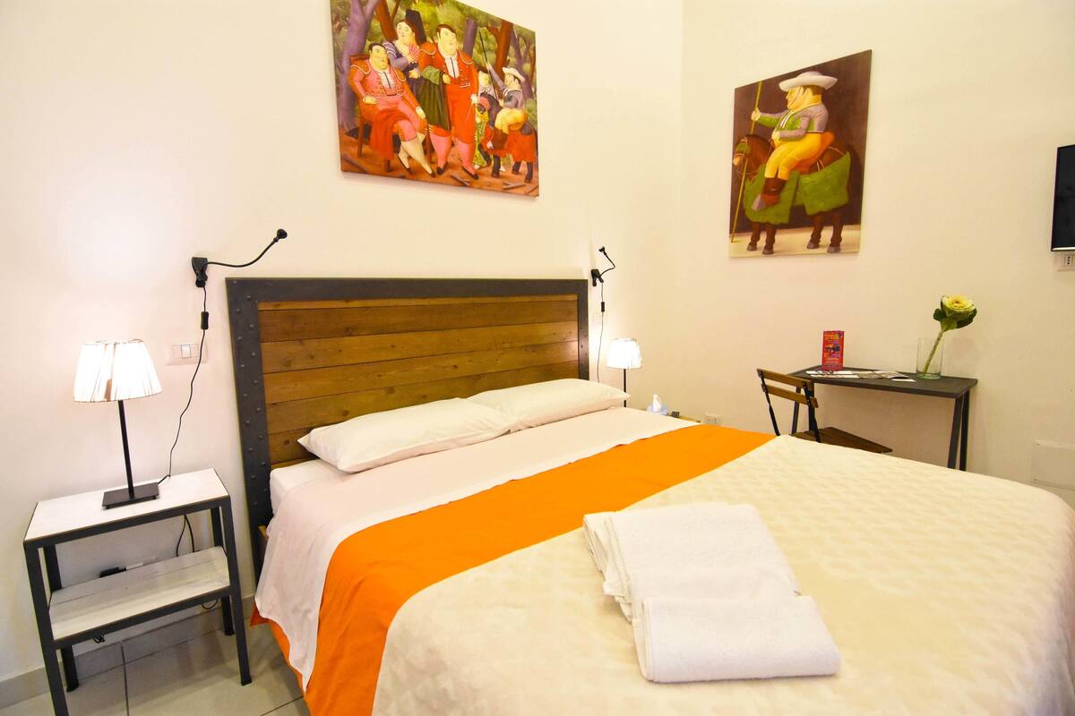 A hotel room with a wooden headboard, a bed with an orange bedspread, a nightstand with a lamp, and two paintings on the wall.