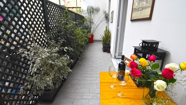 En smal udendørs gang med et træbord, plantekasser og en dekorativ vase med blomster.