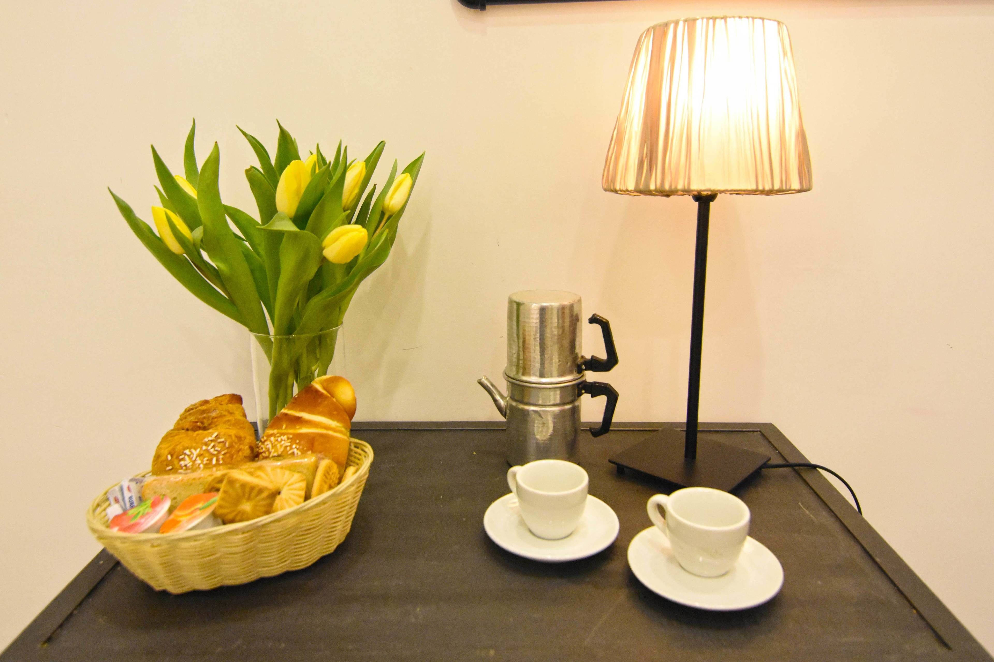 A table with a coffee pot, two cups, a basket of bread, and a vase of tulips.