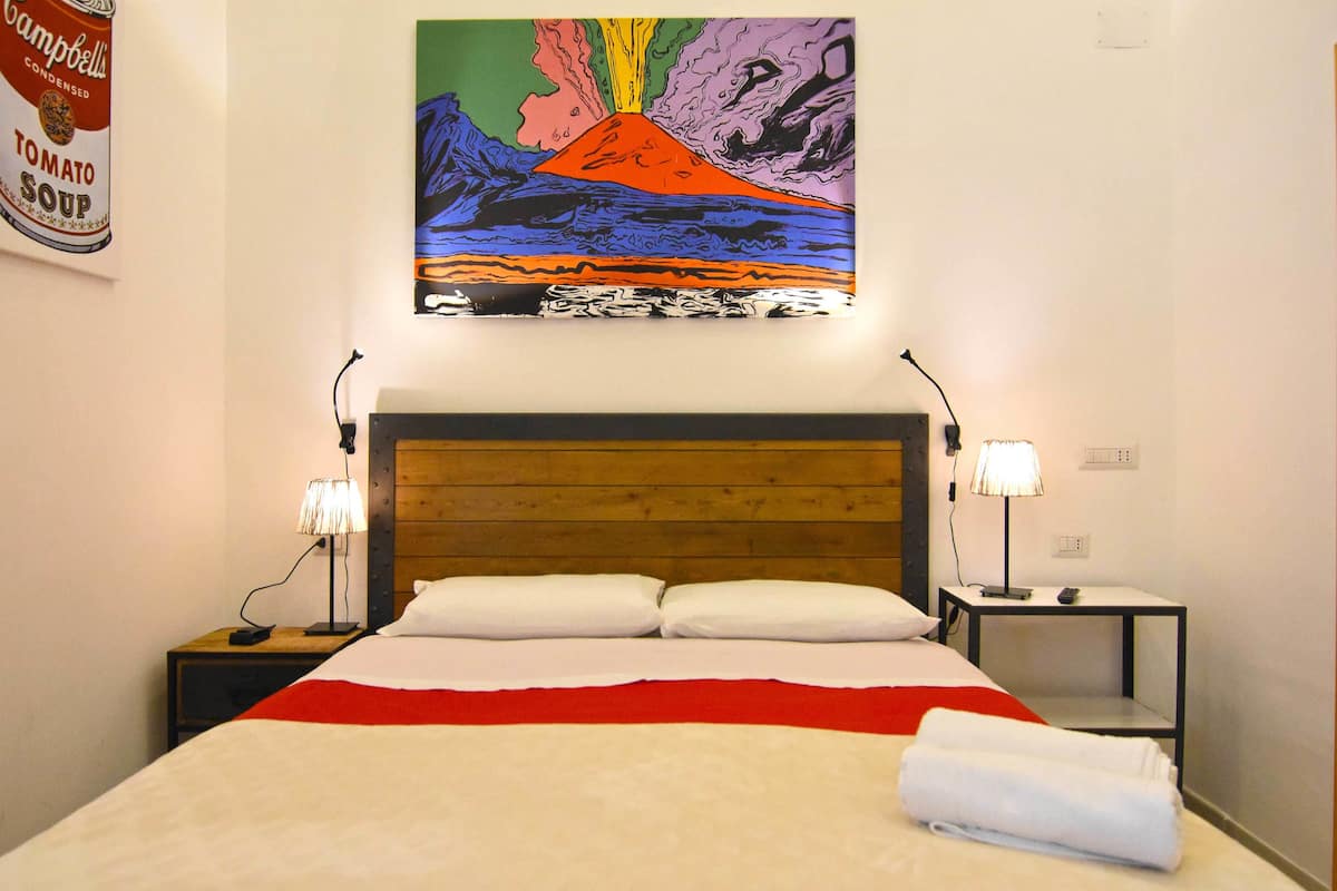 A hotel room with a wooden headboard, a bed with white and red bedding, two bedside tables with lamps, and a colorful painting of a volcano on the wall.