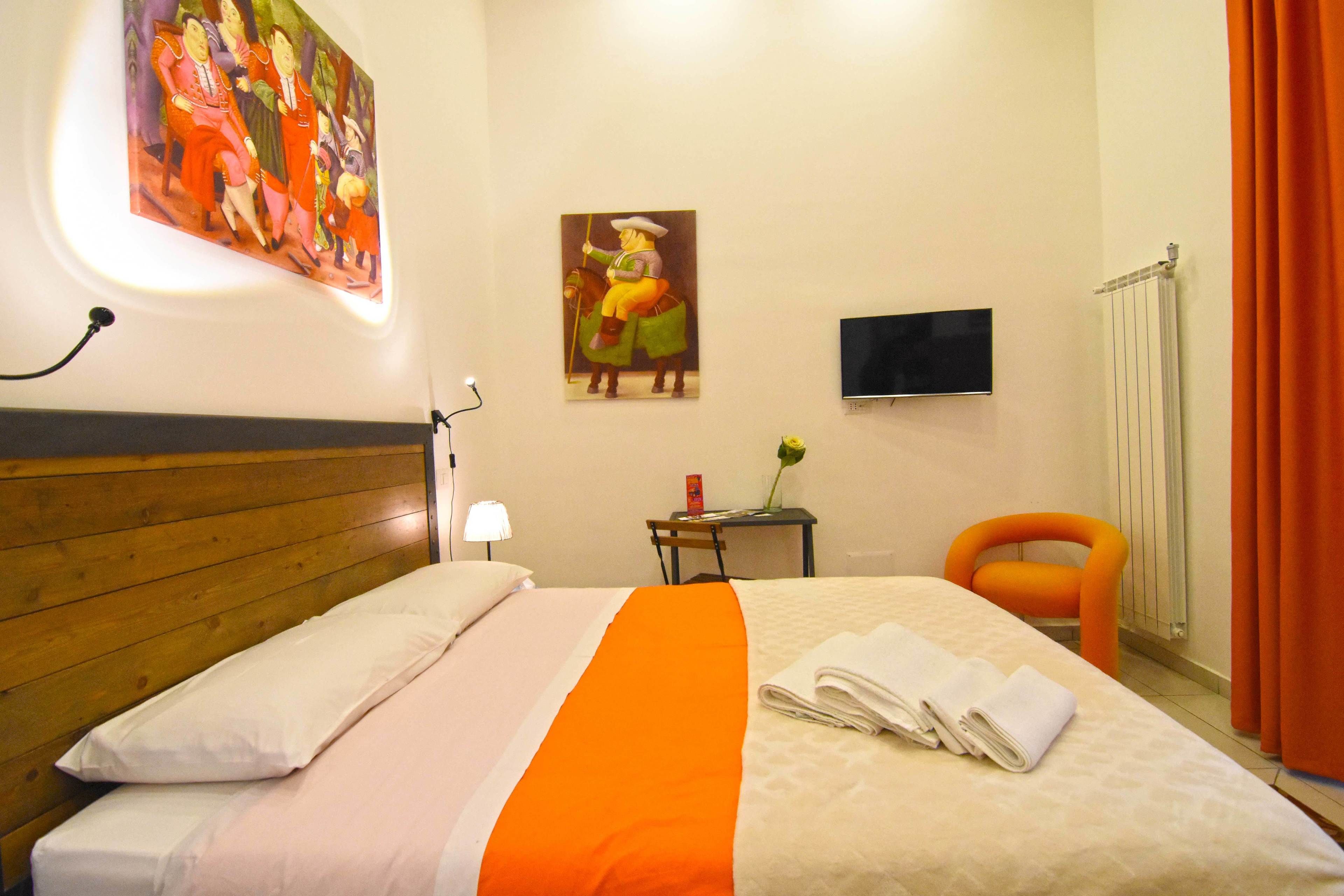 A hotel room with a wooden headboard, a bed with white and orange bedding, a painting on the wall, a small table with a lamp, a chair, and a TV mounted on the wall.