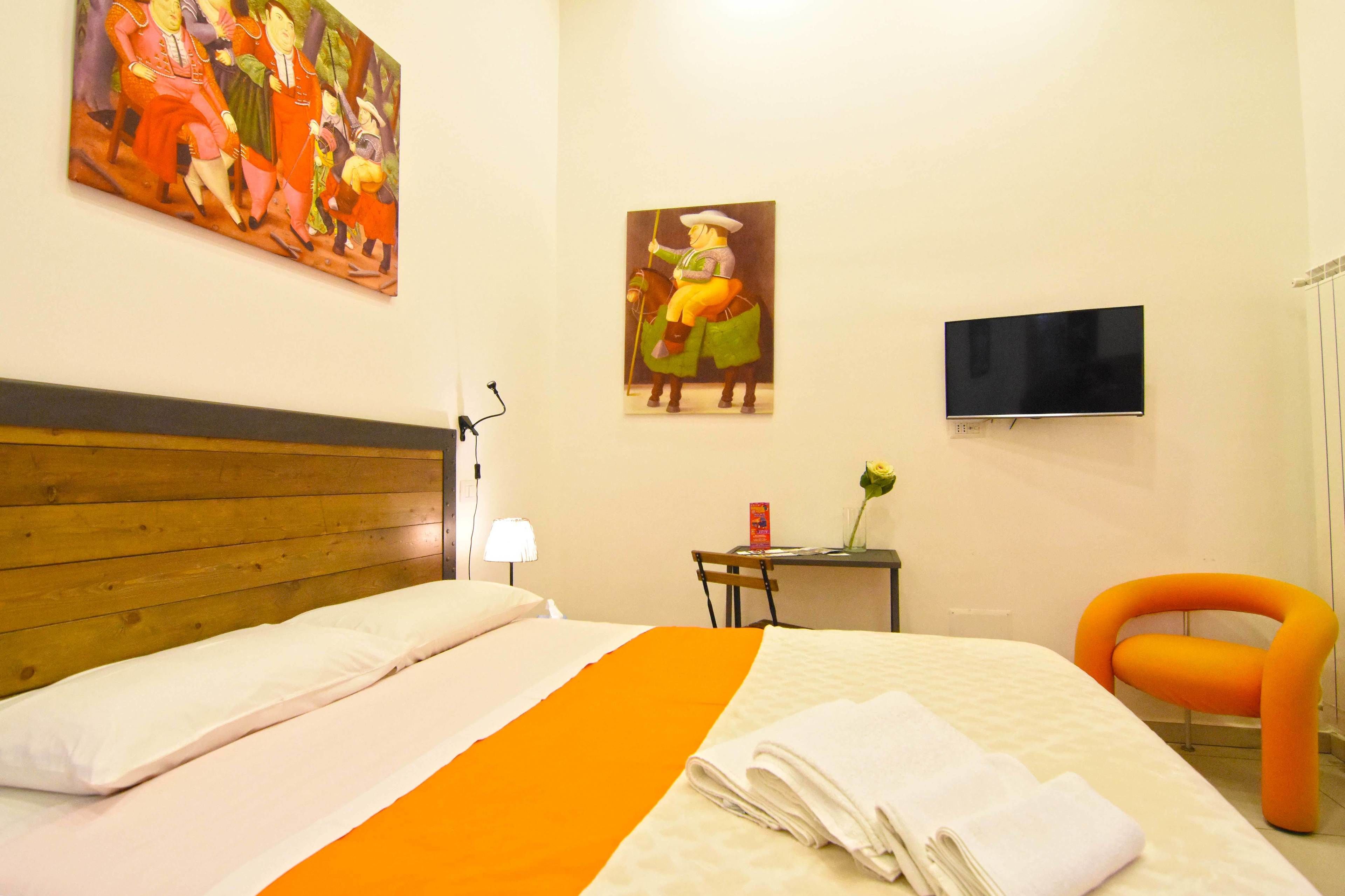 A hotel room with a wooden headboard, a bed with white and orange bedding, a TV mounted on the wall, and a painting of a man on a horse.