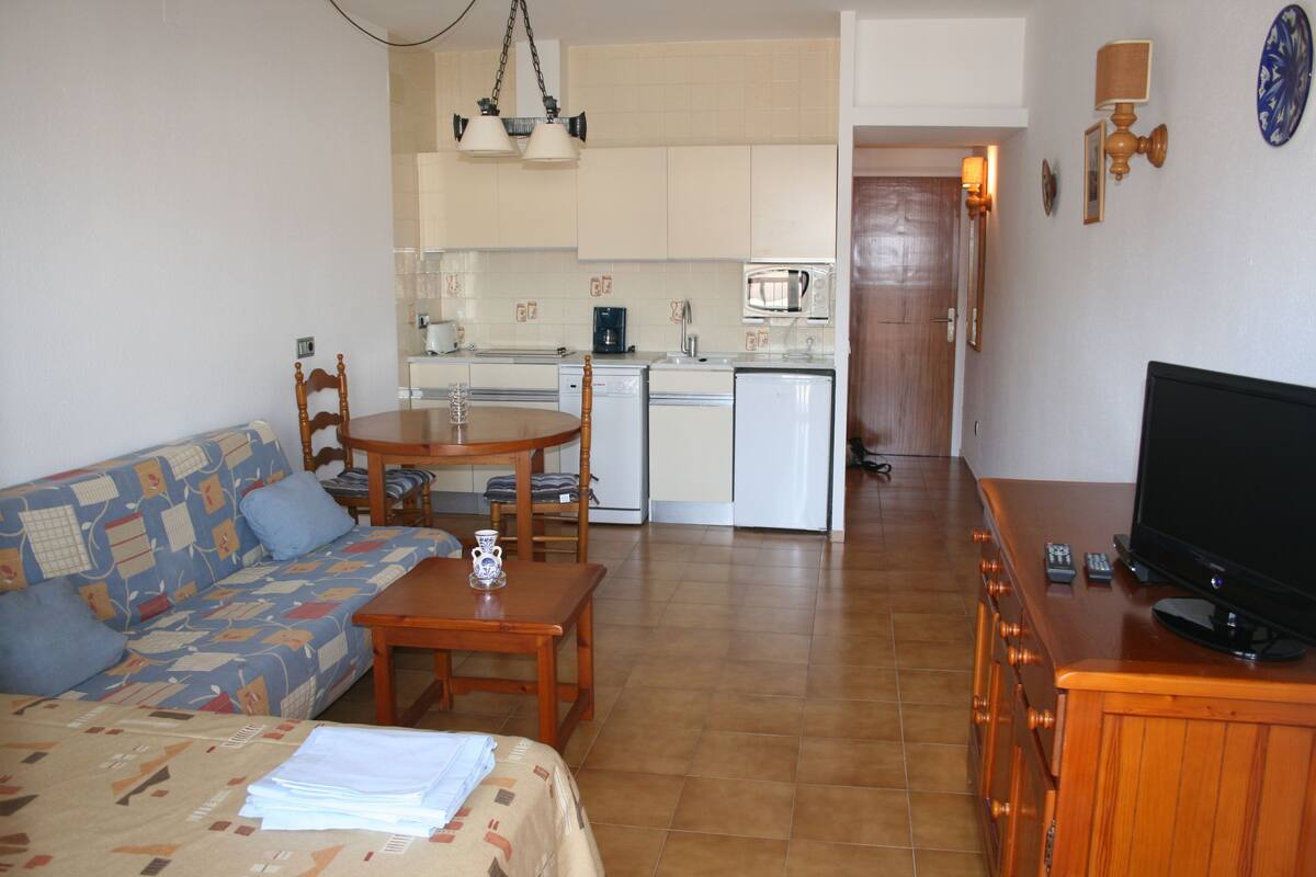 A living room with a sofa, a coffee table, and a TV. There is a kitchen in the background with a microwave, a coffee maker, and a sink.