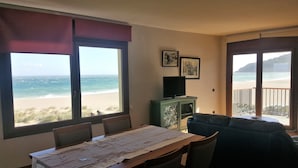 A room with a large window overlooking a beach, a dining table, a sofa, and a television on a cabinet. - Apartaments Les Dunes (Pals)