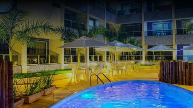 A swimming pool area with lounge chairs and umbrellas, surrounded by a building with multiple floors and windows. - Happy Hotel Ponta Negra (Natal)