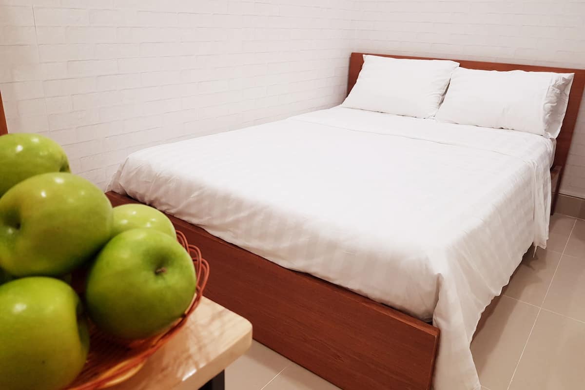 Una cama bien tendida con sábanas blancas y un cabecero de madera, una mesita de noche de madera con una cesta de manzanas verdes, y una pared de ladrillo blanco al fondo.