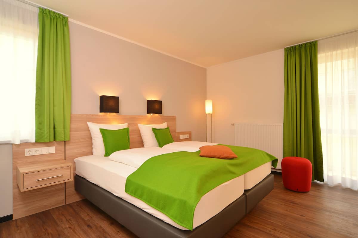 A modern hotel room with a large bed, green curtains, and a red ottoman.