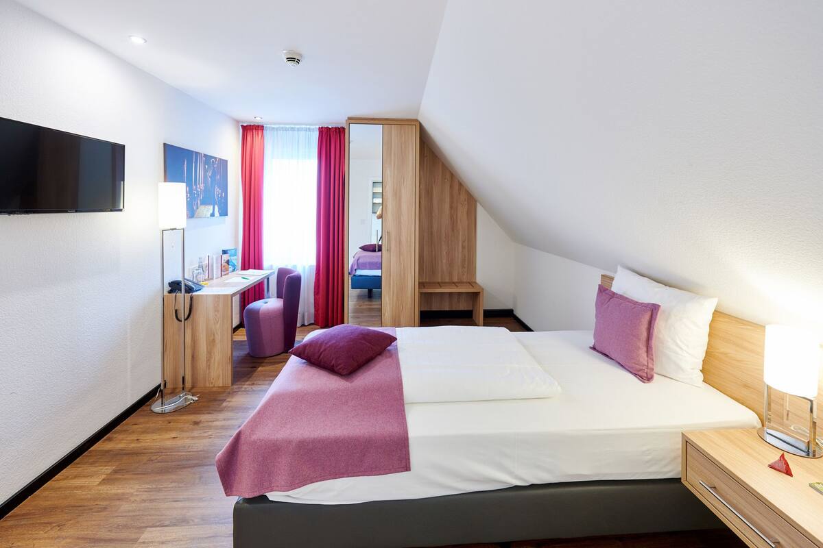 A modern hotel room with a large bed, a desk, a chair, and a TV.