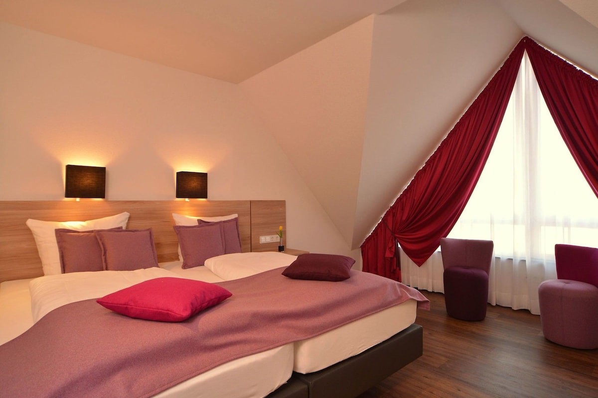 A modern hotel room with a large bed, two bedside lamps, a red curtain, and a seating area with two chairs.