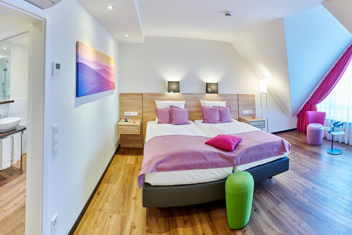 A modern hotel room with a large bed, a sitting area with a sofa and armchair, a small table, and a large window with curtains.
