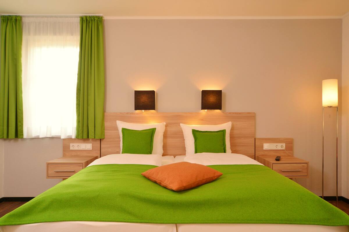 A hotel room with a bed, two bedside tables, a nightstand, a lamp, and a window with green curtains.