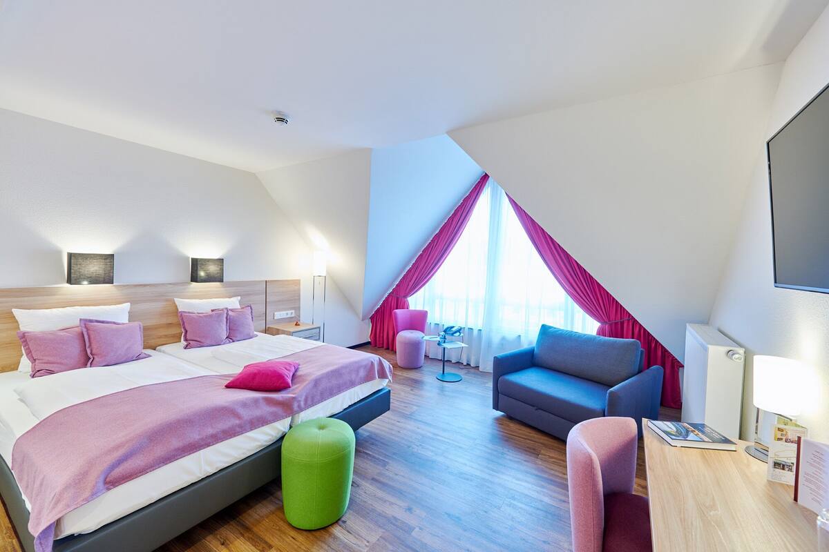A modern hotel room with a large bed, a green ottoman, a desk, and a chair.